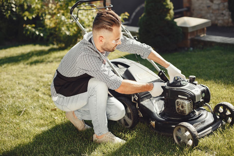 A man wearing protective glasses, a checkered shirt, and light-colored jeans is kneeling on a well-maintained grassy lawn, operating a black petrol-powered lawn mower with a visible engine and control