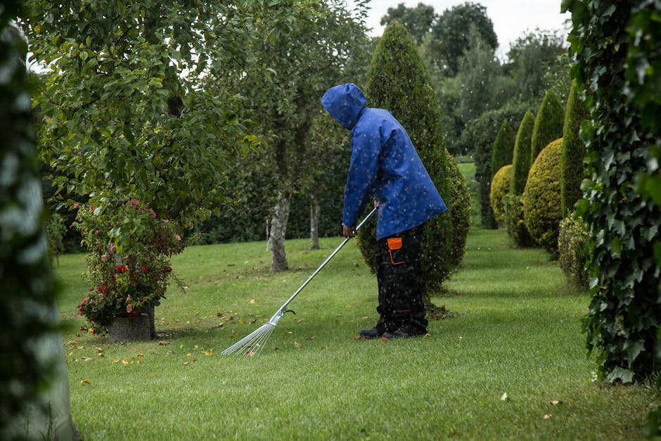 A man wearing protective glasses, a checkered shirt, and light-colored jeans is kneeling on a well-maintained grassy lawn, operating a black petrol-powered lawn mower with a visible engine and control